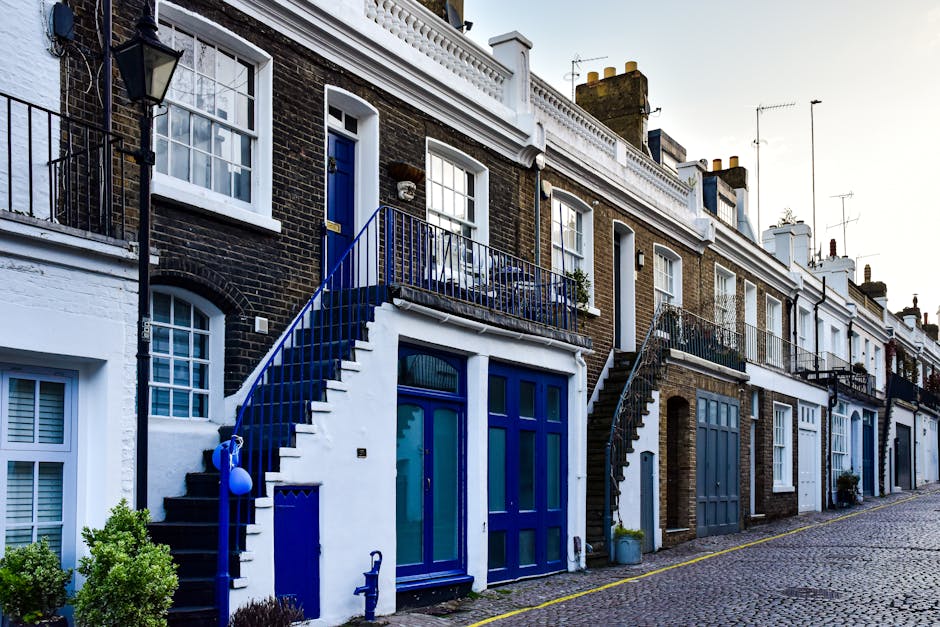 A row of terraced houses on Becontree Estate with brick facades and white window frames, featuring external staircase entrances with black and blue railings leading to first-floor doors. The ground floors have large windows, some with curtains, and blue-painted doors. There are small potted plants and bushes near the entrances. The street is paved with cobblestones, with a narrow yellow line along the curb. The scene is captured during daylight hours with clear weather, highlighting the residential environment typical of property relocations and house moving services. Man With a Van Dagenham frequently handles furniture transport and home relocation projects, including packing, loading, and stairway access in such buildings.
