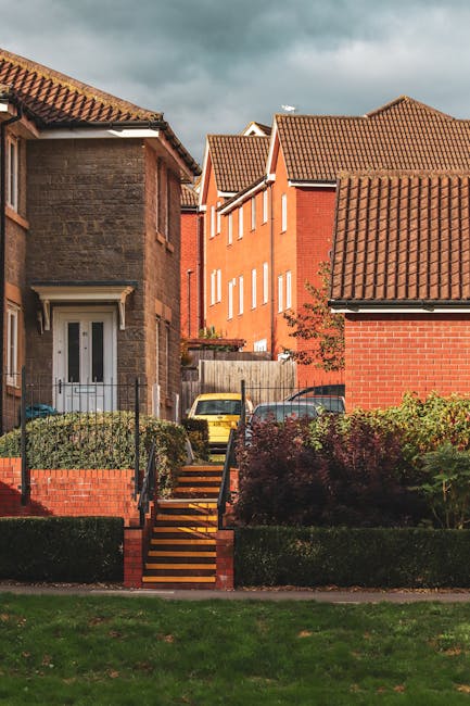 A row of terraced houses on Becontree Estate with brick facades and white window frames, featuring external staircase entrances with black and blue railings leading to first-floor doors. The ground floors have large windows, some with curtains, and blue-painted doors. There are small potted plants and bushes near the entrances. The street is paved with cobblestones, with a narrow yellow line along the curb. The scene is captured during daylight hours with clear weather, highlighting the residential environment typical of property relocations and house moving services. Man With a Van Dagenham frequently handles furniture transport and home relocation projects, including packing, loading, and stairway access in such buildings.