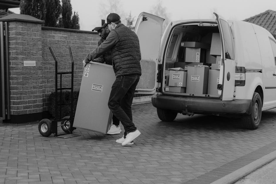 Two men are engaged in a home relocation process, seen on a paved driveway next to a brick wall. One man, wearing a cap, glasses, a padded jacket, and jeans, is lifting a large cardboard box with a fragile label, while the other man, dressed in a baseball cap, dark jacket, and sneakers, assists by stabilizing the box. They are transferring packed boxes from a white moving van parked nearby with its rear doors open, revealing more boxes inside secured on the floor. A two-wheeled hand trolley stands nearby, used for transporting heavier items, which appears to be in preparation for furniture transport or packing and moving activities. The scene is outdoors, with natural lighting, and the van is positioned adjacent to the property, indicating an ongoing load or unload process during a house removal handled by Man With a Van Dagenham, capturing the logistics involved in residential moves and furniture transportation.