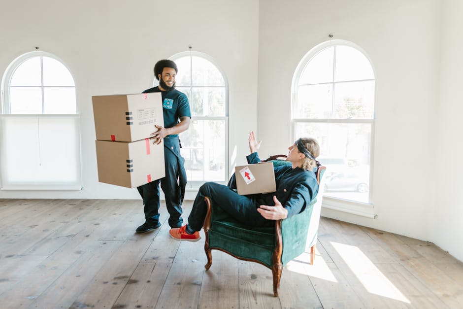 A professional mover from Man With a Van Dagenham is carrying two medium-sized cardboard boxes stacked one on top of the other, heading towards an open doorway inside a bright, spacious room with wood