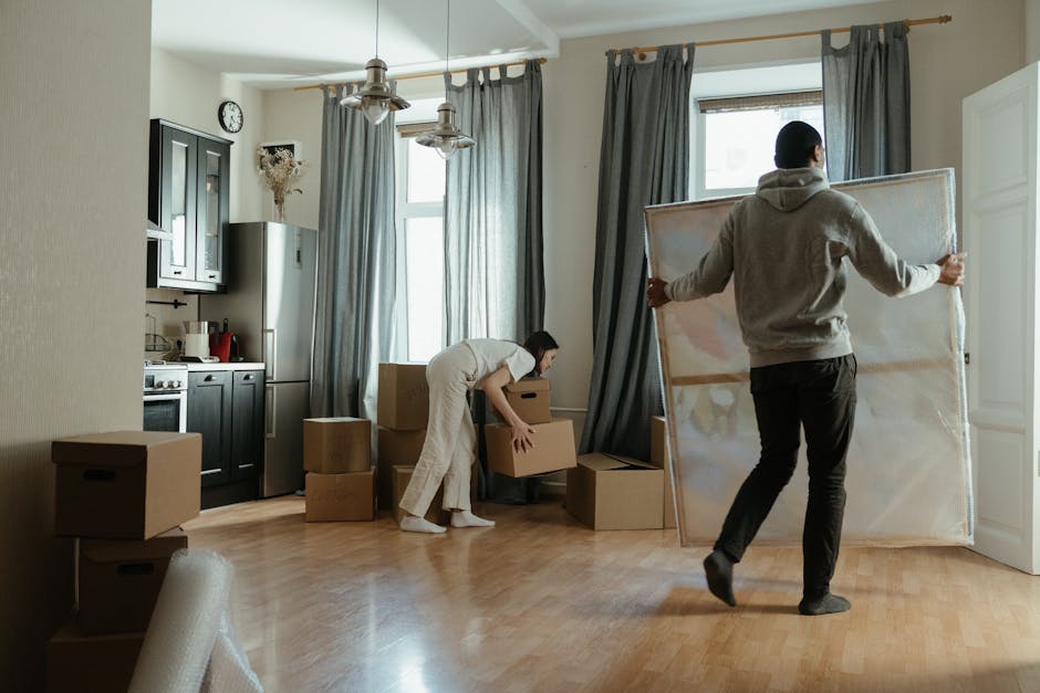 A professional mover from Man With a Van Dagenham is carrying two medium-sized cardboard boxes stacked one on top of the other, heading towards an open doorway inside a bright, spacious room with wood