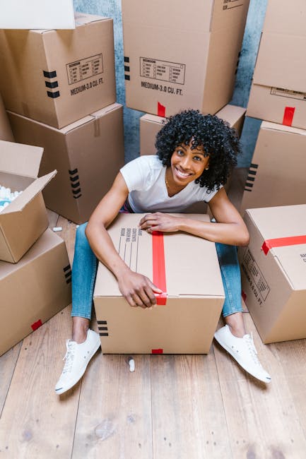 A young woman with curly black hair and a bright smile is seated on a wooden floor surrounded by numerous neatly packed cardboard boxes, some sealed with red tape and labeled with storage information. She is wearing a white T-shirt, blue jeans, and white sneakers. The setting appears to be inside a property, possibly a room or hallway, with blue-painted walls visible in the background. Several boxes are stacked in the environment, indicating an active packing or home relocation process. The woman is holding a large box on her lap, suggesting involvement in furniture transport or packing during a house move. This scene exemplifies packing and moving logistics handled by Man With a Van Dagenham, a professional removals service specializing in home removals and transport solutions, as part of a comprehensive home relocation or furniture transport service.