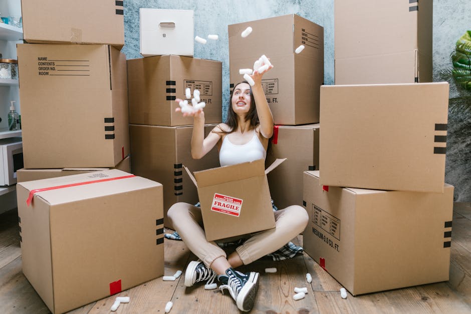 A woman sitting on a wooden floor surrounded by packed cardboard boxes of various sizes, some labeled with red and black tape, inside a room with grey walls and a small white cupboard in the background. She is smiling and throwing packing peanuts into the air while sitting cross-legged with an open box in front of her. The scene depicts the process of packing for a home relocation, with protective packaging materials and furniture boxes ready for transport. A green plant is visible on the right side of the image, and ambient lighting illuminates the interior. This setting illustrates a professional moving service, such as those provided by Man With a Van Dagenham, preparing items for furniture transport and house removals, aligning with the topic of same-day removals availability and moving logistics in Dagenham.