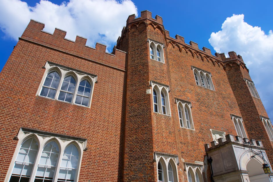 A close-up view of a large historic brick building with tall, arched white-framed windows and decorative battlements along the roofline, set against a bright blue sky with scattered white clouds. The building's exterior features red bricks with some variations in tone, and a small stone archway entrance with a lantern hanging beside it. This architectural style is indicative of a castle or historic structure, often associated with museums or heritage sites. The scene is well-lit with natural daylight, highlighting the architectural details and the textures of the brickwork. In the context of house removals and relocation services by Man With a Van Dagenham, this image could represent the preparation for moving artifacts, furniture, or equipment related to a historical or museum area, emphasizing careful handling and transport during the home relocation process for historic properties.