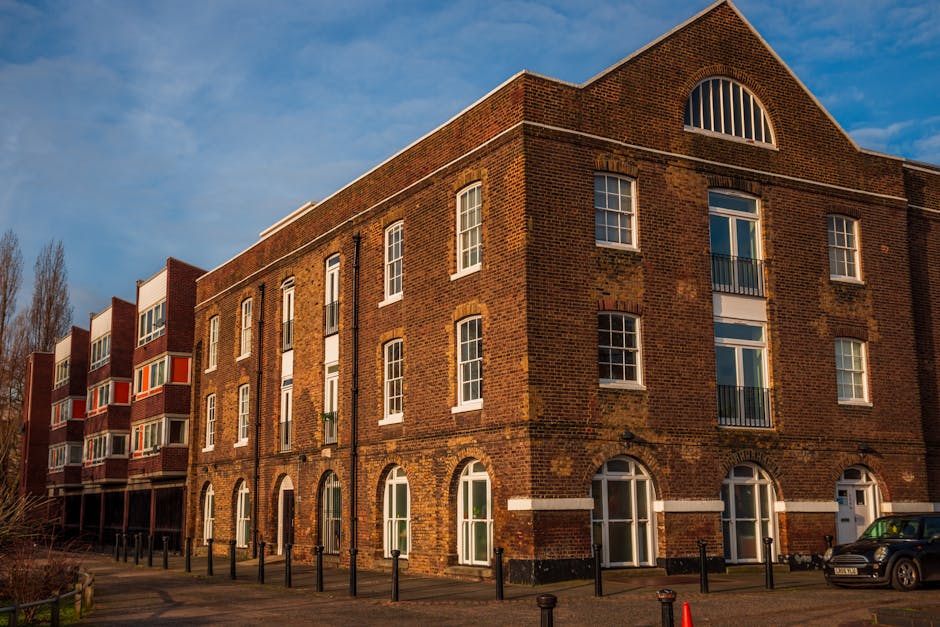 A close-up view of a large historic brick building with tall, arched white-framed windows and decorative battlements along the roofline, set against a bright blue sky with scattered white clouds. The building's exterior features red bricks with some variations in tone, and a small stone archway entrance with a lantern hanging beside it. This architectural style is indicative of a castle or historic structure, often associated with museums or heritage sites. The scene is well-lit with natural daylight, highlighting the architectural details and the textures of the brickwork. In the context of house removals and relocation services by Man With a Van Dagenham, this image could represent the preparation for moving artifacts, furniture, or equipment related to a historical or museum area, emphasizing careful handling and transport during the home relocation process for historic properties.
